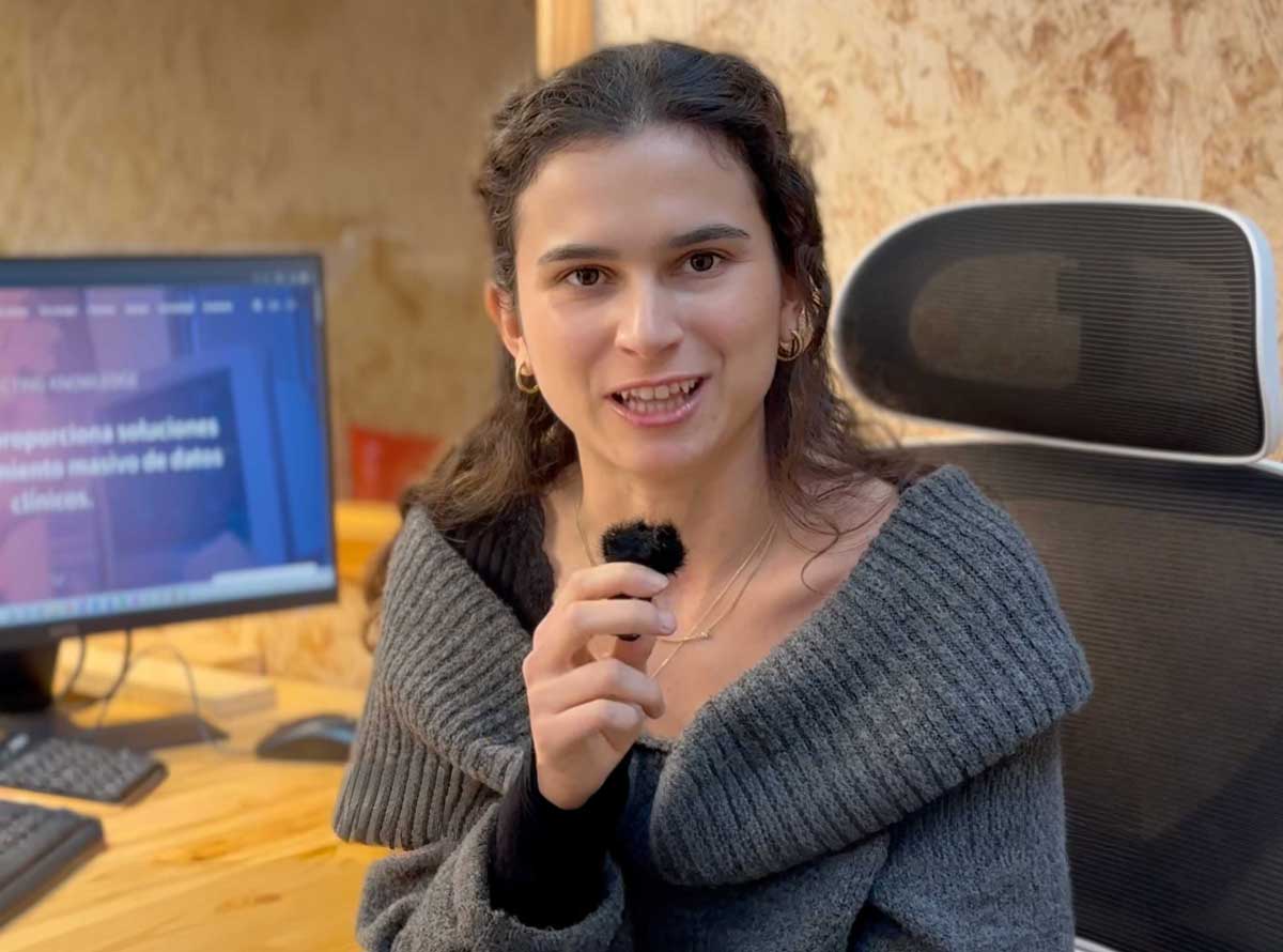 Una mujer joven con suéter gris oscila sosteniendo un micrófono pequeño en una oficina con escritorio y monitor al fondo.