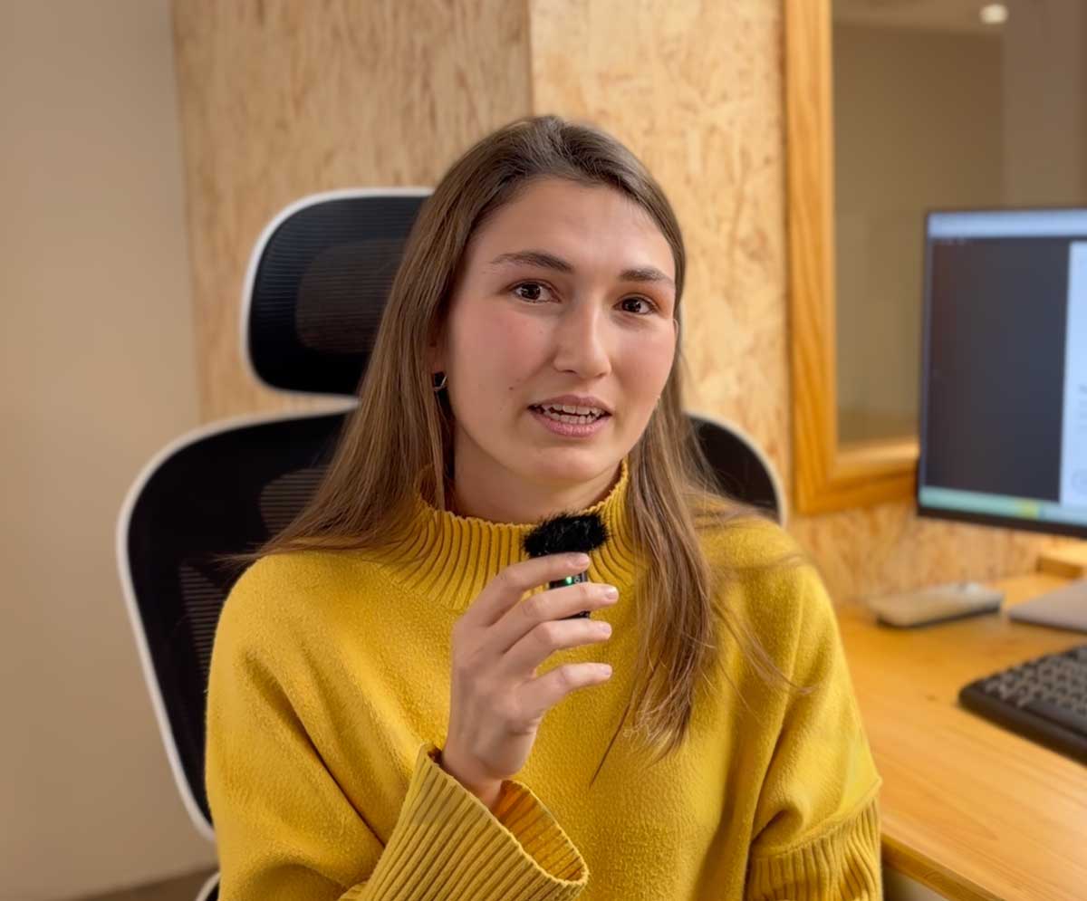 Mujer con suéter amarillo sosteniendo un micrófono pequeño en una oficina con escritorio y computadora.