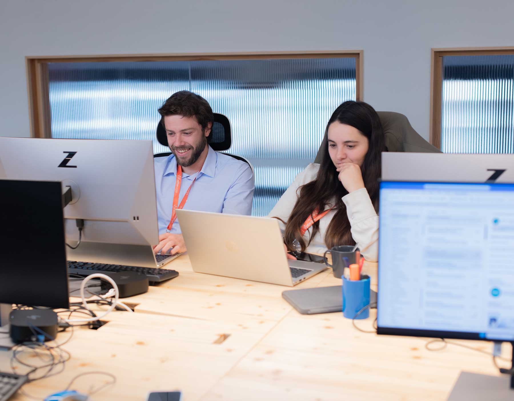Dos personas trabajando juntas en una oficina, una sonríe mientras usa un ordenador portátil y la otra mira pensativa su pantalla.