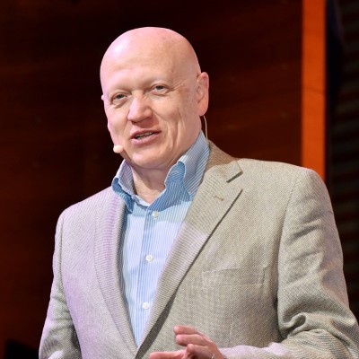 Hombre calvo con chaqueta gris y camisa azul claro hablando con un micrófono de diadema en un escenario.