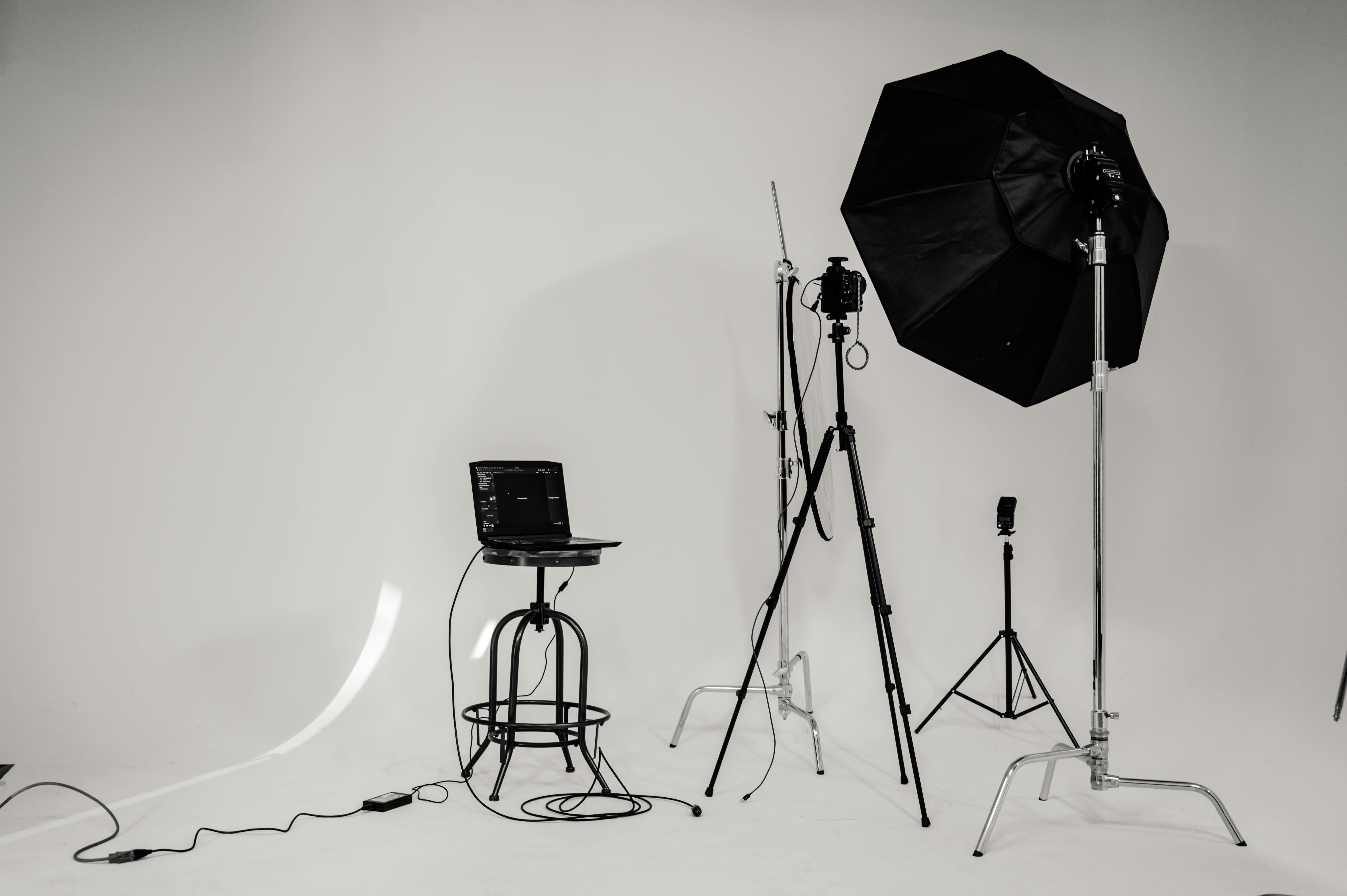 Photography studio setup with a laptop on a stool, camera on a tripod, and lighting equipment with large softbox and flash on stands against a white backdrop.