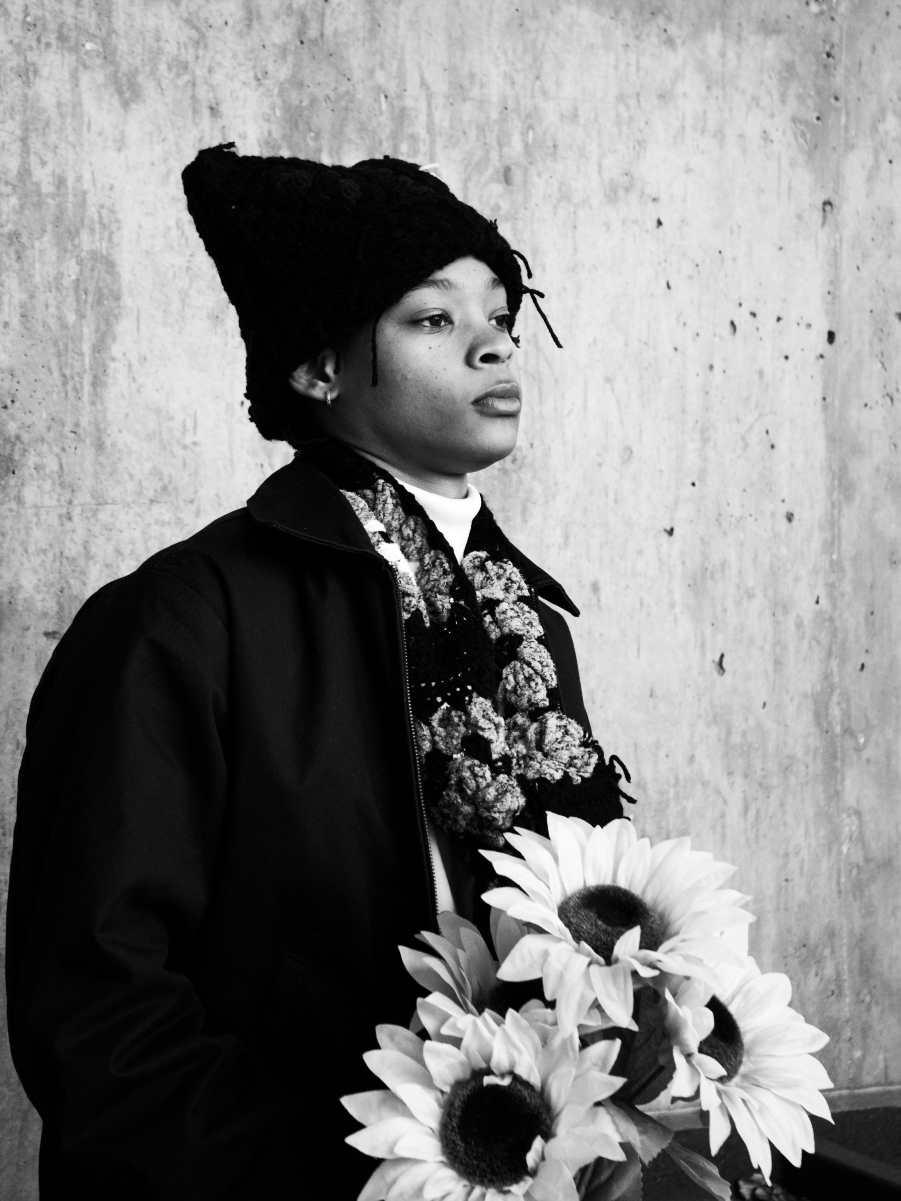 Black and white photo of a person wearing a knitted hat and scarf, holding a bouquet of three large sunflowers, standing against a concrete wall.