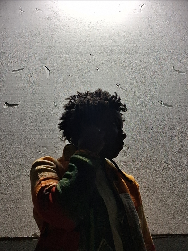 Silhouette of a person with curly hair wearing a colorful jacket against a textured wall lit from above.