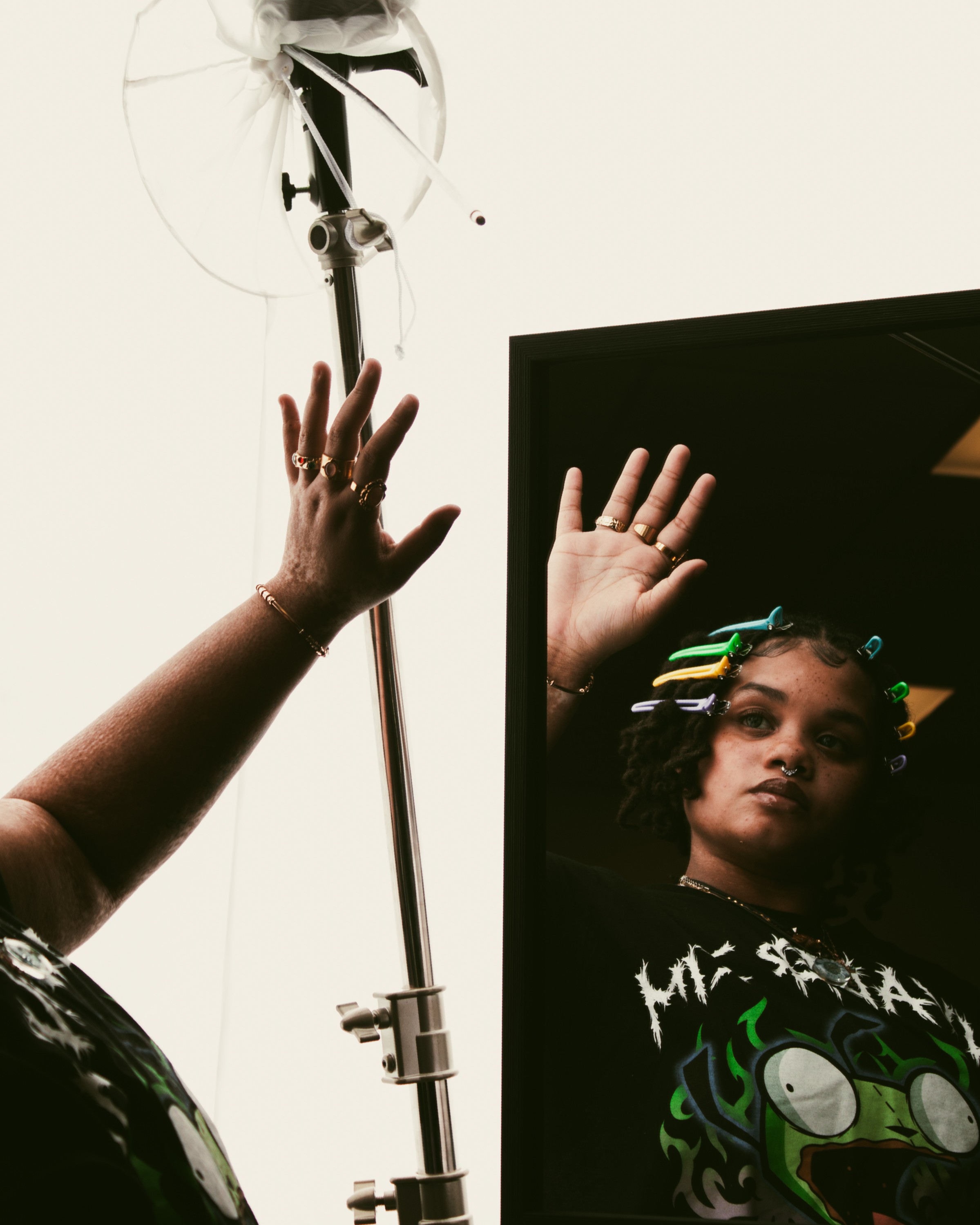 Person with colorful hair clips looks at their reflection in a mirror while raising their hand.