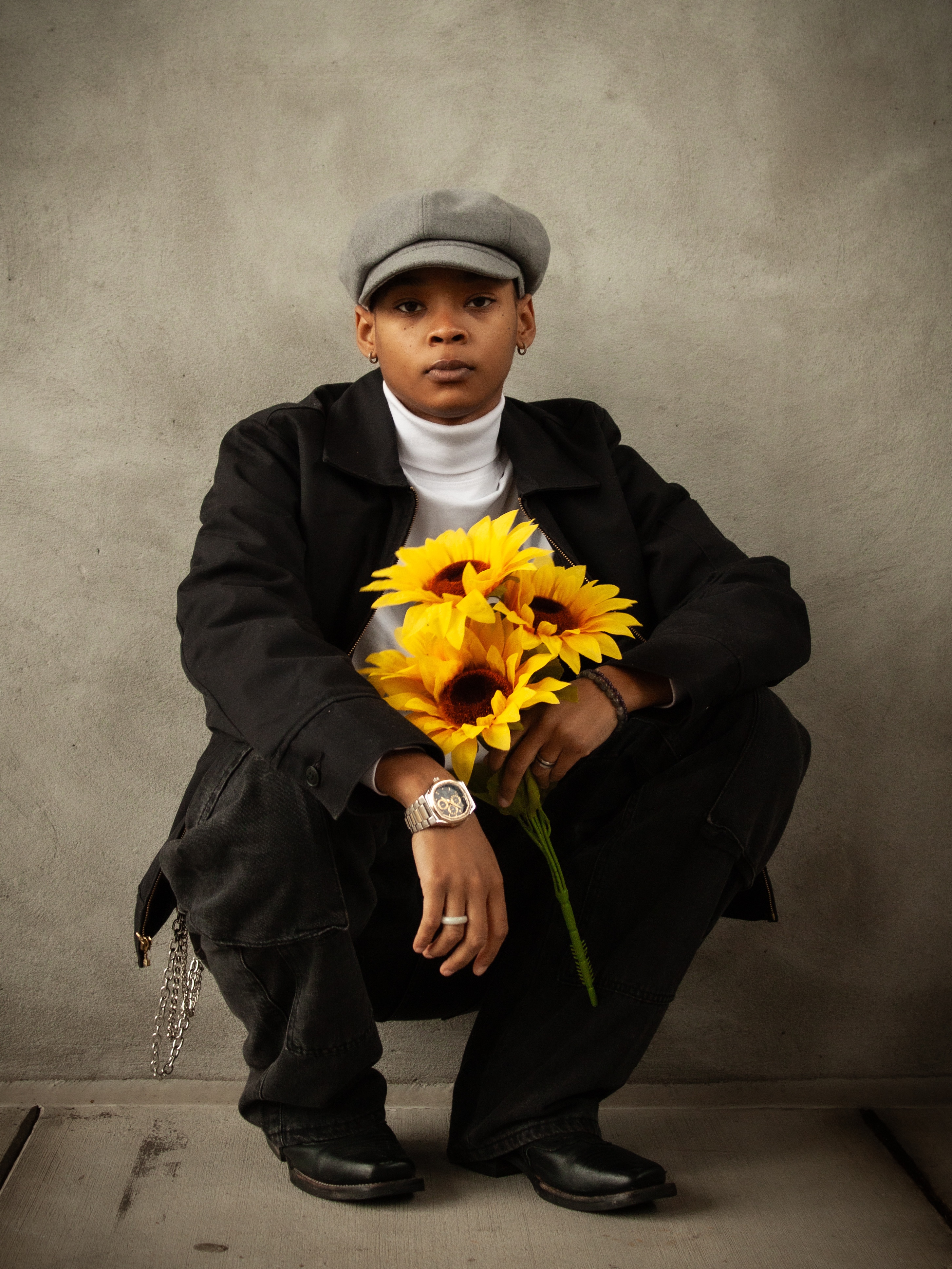 Person squatting against a gray wall holding a bouquet of bright yellow sunflowers.