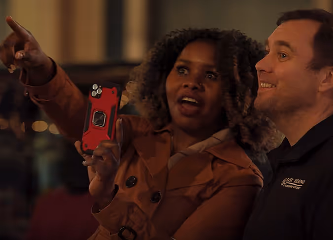 Woman holding a red smartphone and pointing at something while a man smiles beside her in a dimly lit setting.