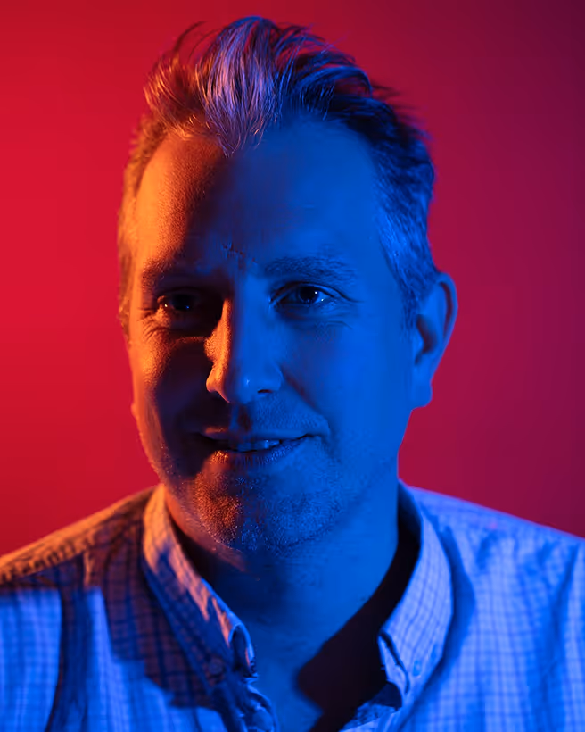 Man with gray hair wearing a checkered shirt, illuminated with blue and orange lighting against a red background.