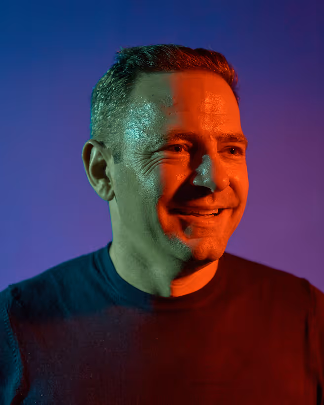 Smiling man with short hair illuminated by green and red lighting against a blue-purple background.