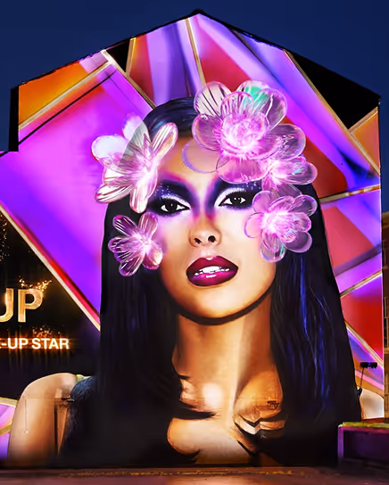 Portrait of a woman with bold purple makeup and translucent pink flowers overlaying her face on a vibrant geometric background.