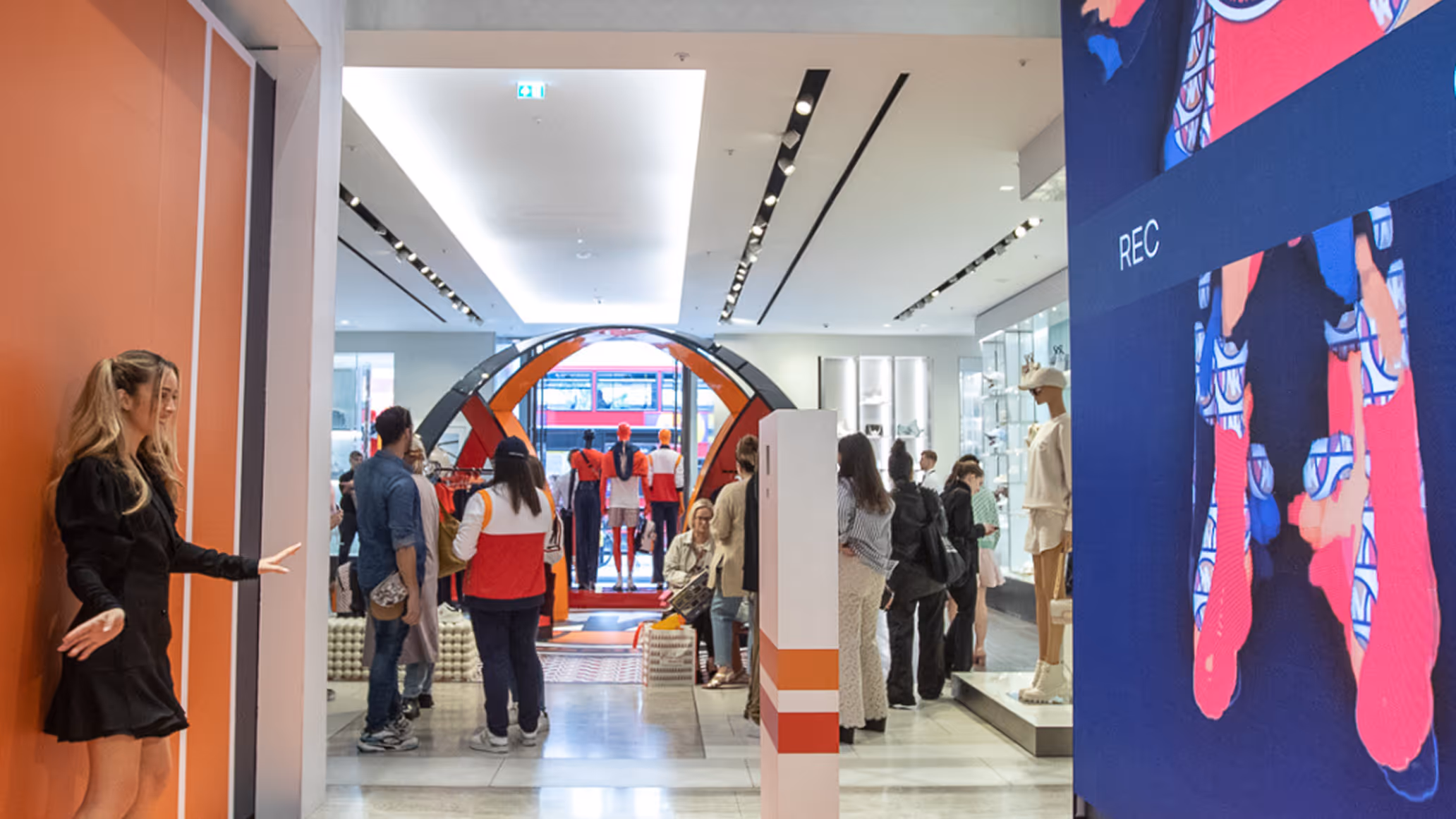 People inside a modern clothing store with mannequins, a digital screen showing a fashion recording, and a woman in black near an orange wall.