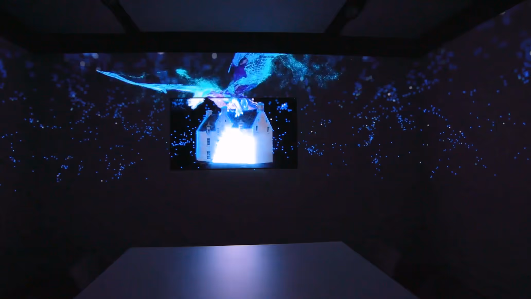 Dark room with blue digital light patterns projecting onto walls and a screen showing a glowing house model.