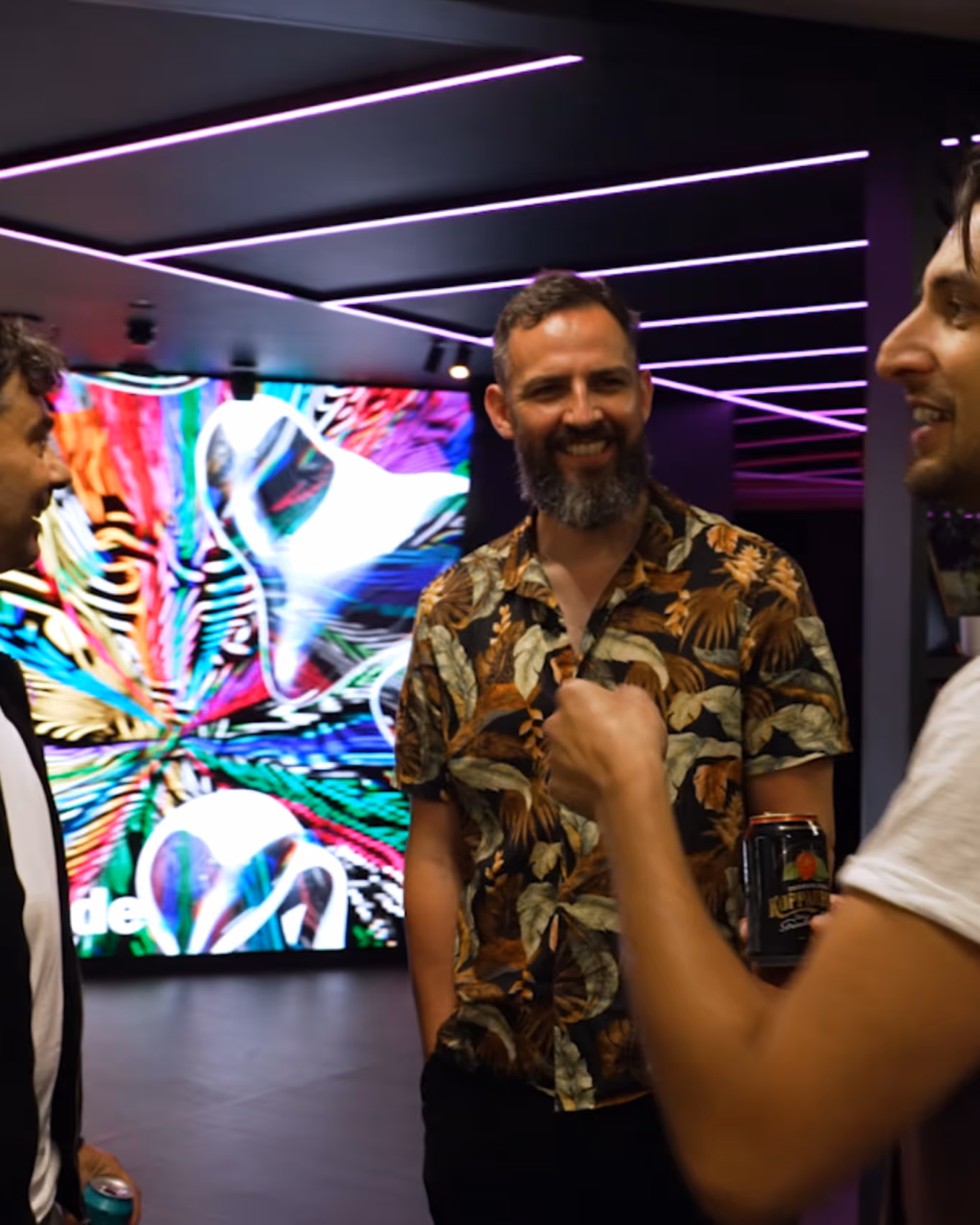 Three men conversing indoors with colorful abstract digital art in the background and purple neon lights overhead.
