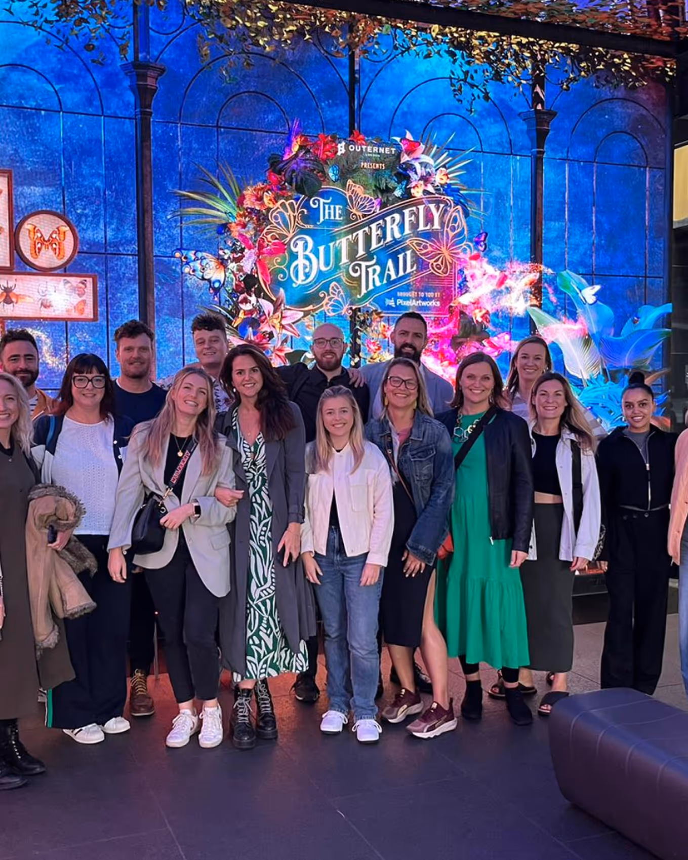 Group of smiling people standing in front of a colorful illuminated sign that reads 'The Butterfly Trail'.