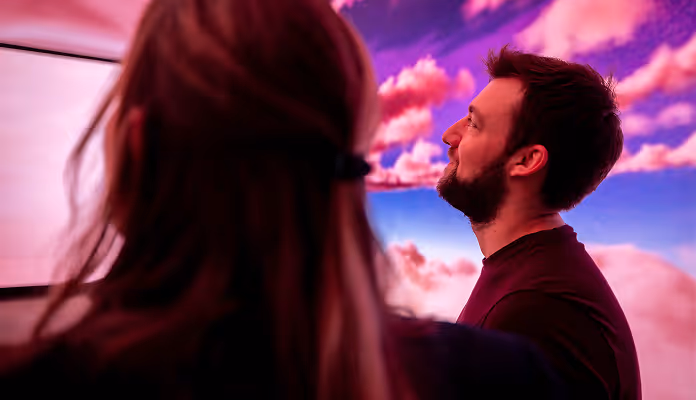 Man with a beard looking up at a large screen displaying a colorful sky with pink clouds, with a person seen from behind in the foreground.