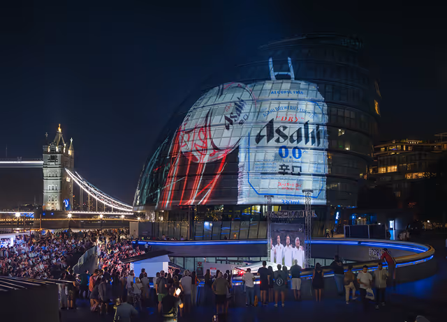 Night view of a crowd watching a large projection of an Asahi 0.0 alcohol-free beer can displayed on a round glass building near a lit Tower Bridge.
