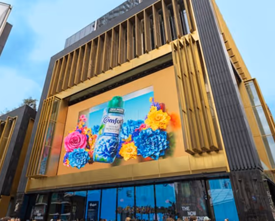 Outdoor building facade with a large digital billboard displaying a Comfort fabric softener bottle surrounded by colorful pink, blue, and yellow flowers.
