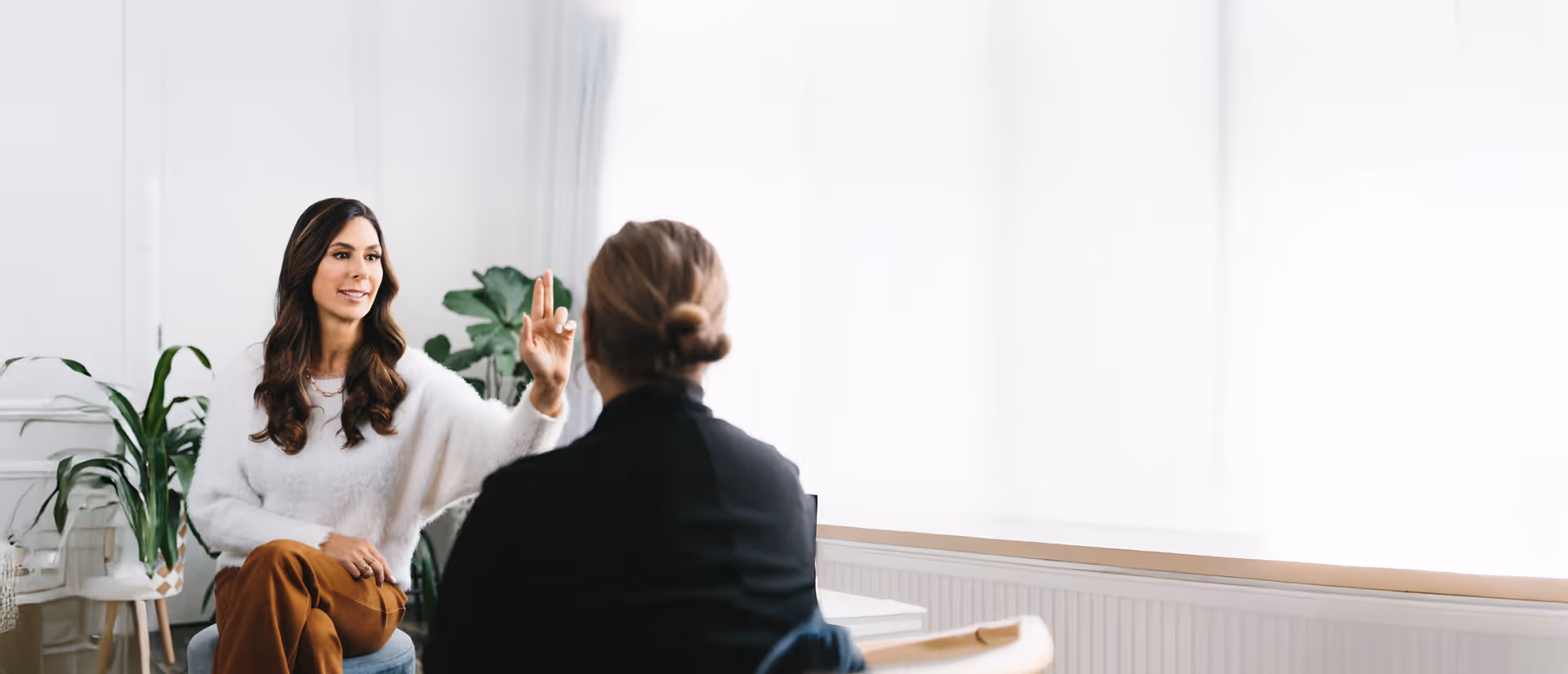 Woman in white sweater gesturing with three fingers while talking to another person in a bright room with plants.