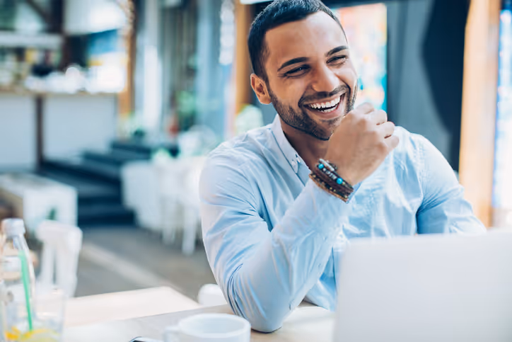 Smiling man with beaded bracelets sitting at a table with a laptop in a bright, modern cafe.