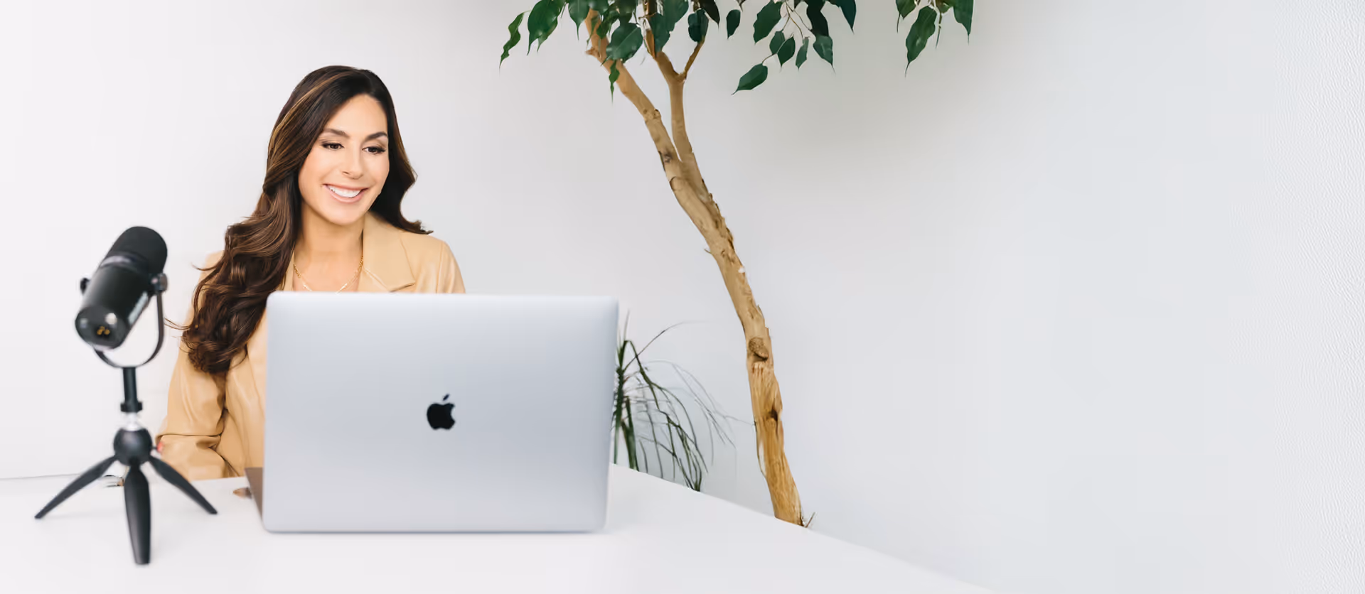 Smiling woman sitting at a white desk with a laptop and microphone for podcast recording.