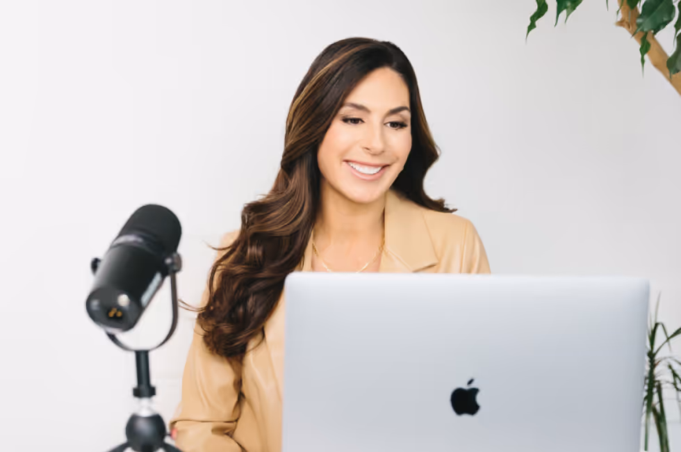 Smiling woman with long hair in beige jacket working on a laptop with a microphone nearby.