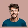 Smiling young man with dark hair and beard wearing a blue shirt against a pink background.