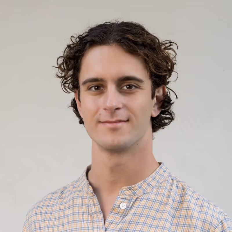 Young man with curly dark hair wearing a light plaid shirt, smiling slightly against a plain light background.