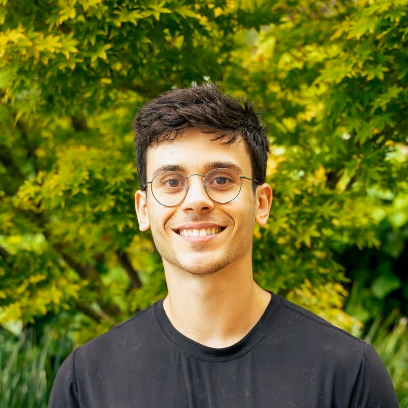 Young man with short dark hair and glasses smiling in front of green leafy background.