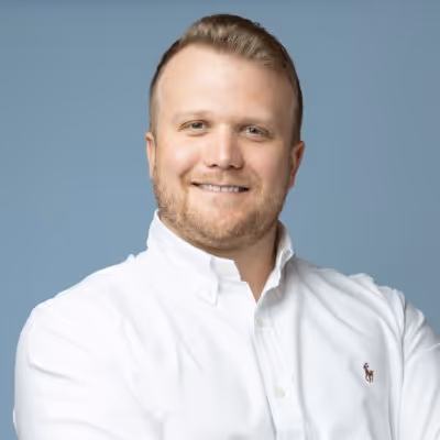 Smiling man with short blond hair and beard wearing a white button-up shirt against a blue background.