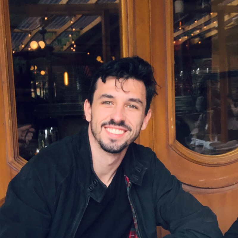 Portrait of a young man with short dark hair and beard wearing a black jacket and white shirt against a white background.
