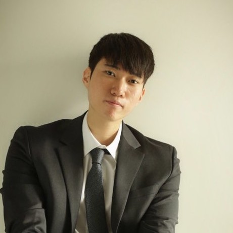 Young man with short curly hair smiling, wearing a dark shirt and a round pendant necklace.
