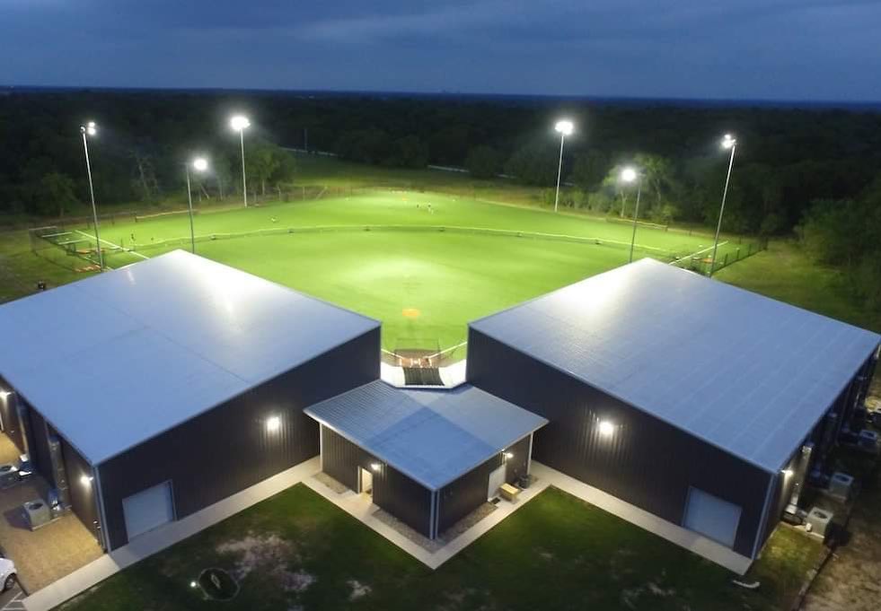 Aerial night view of a well-lit baseball field with two large adjacent buildings in the foreground.