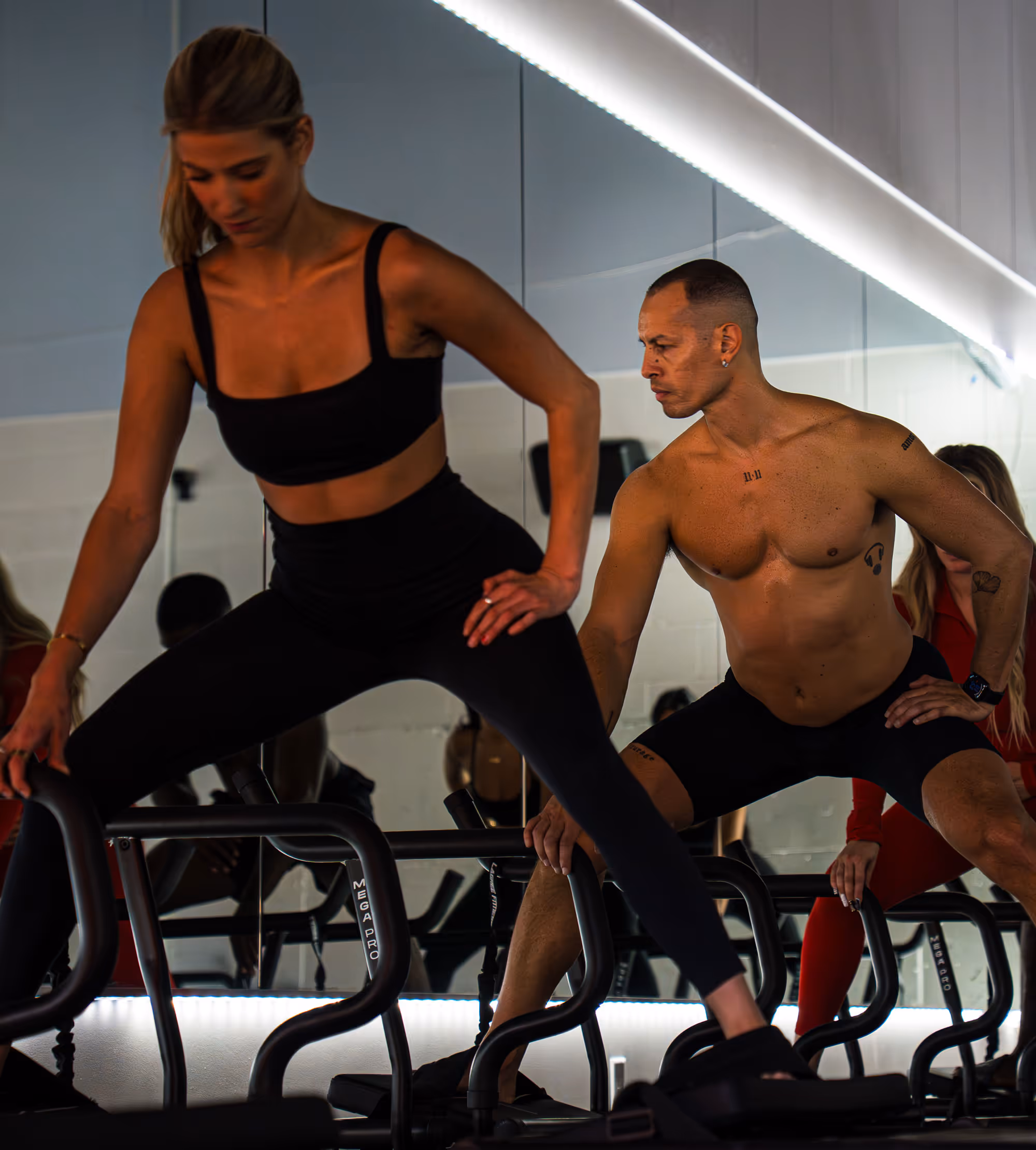 Two individuals performing a side stretch on black MegaPro fitness equipment in a gym with mirrored walls.
