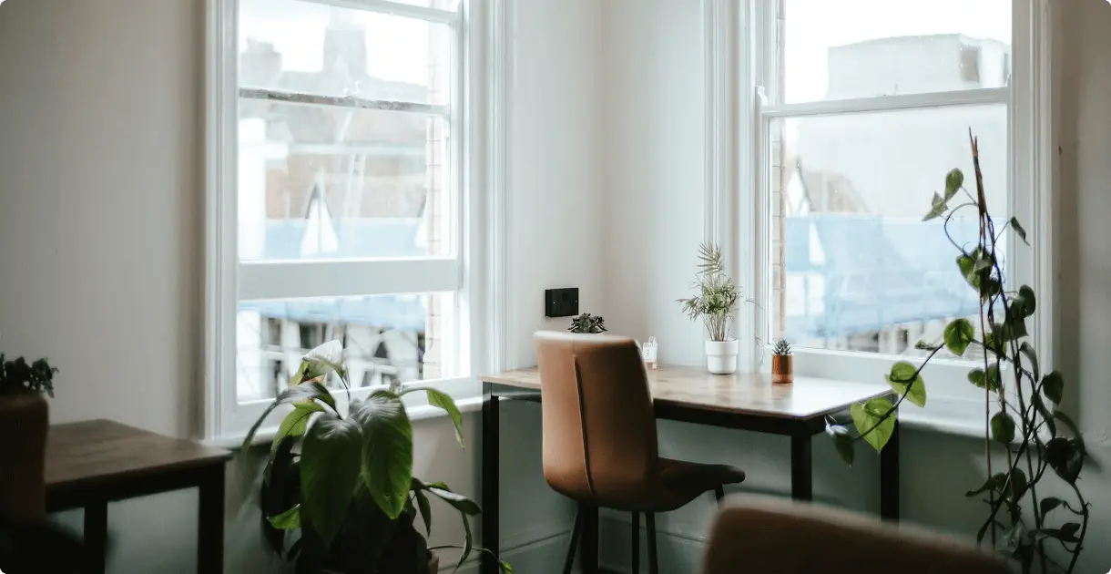 Gemütlicher, minimalistischer Arbeitsbereich mit einem Holztisch, einem braunen Stuhl und Zimmerpflanzen in der Nähe großer, heller Fenster.