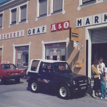 Small group of people standing near a dark Suzuki SUV and a red car in front of a building with signs reading GRAF A&O MARKT.