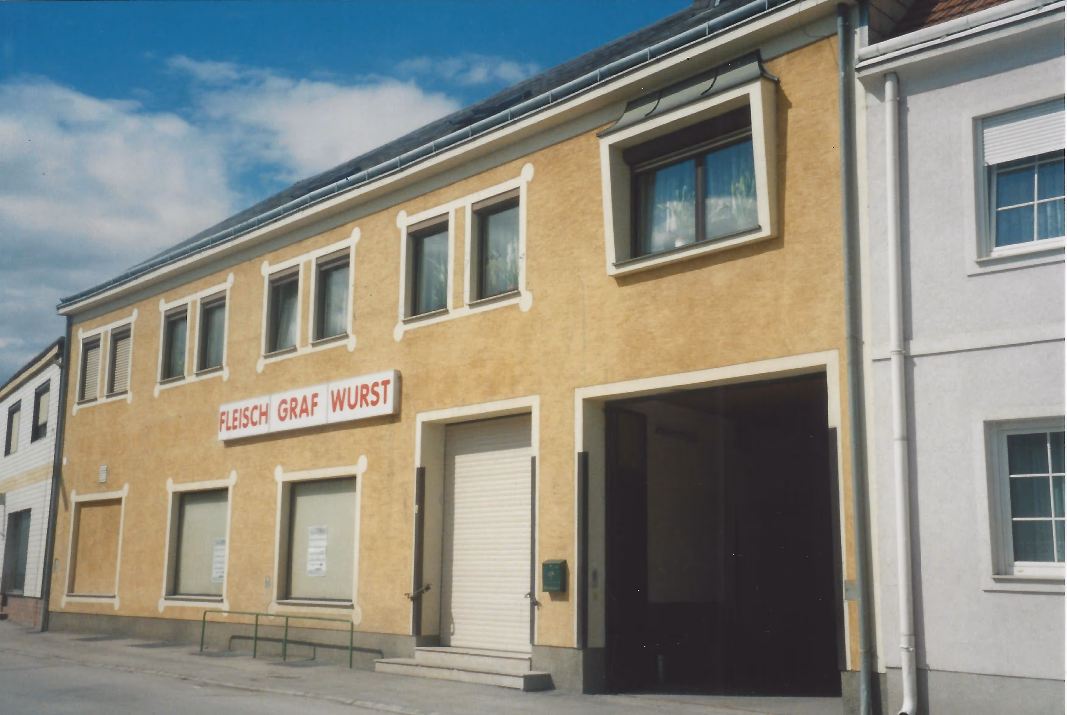 Yellow building with multiple windows and a sign reading 'Fleisch Graf Wurst' above a closed white roller door and a large open garage door.