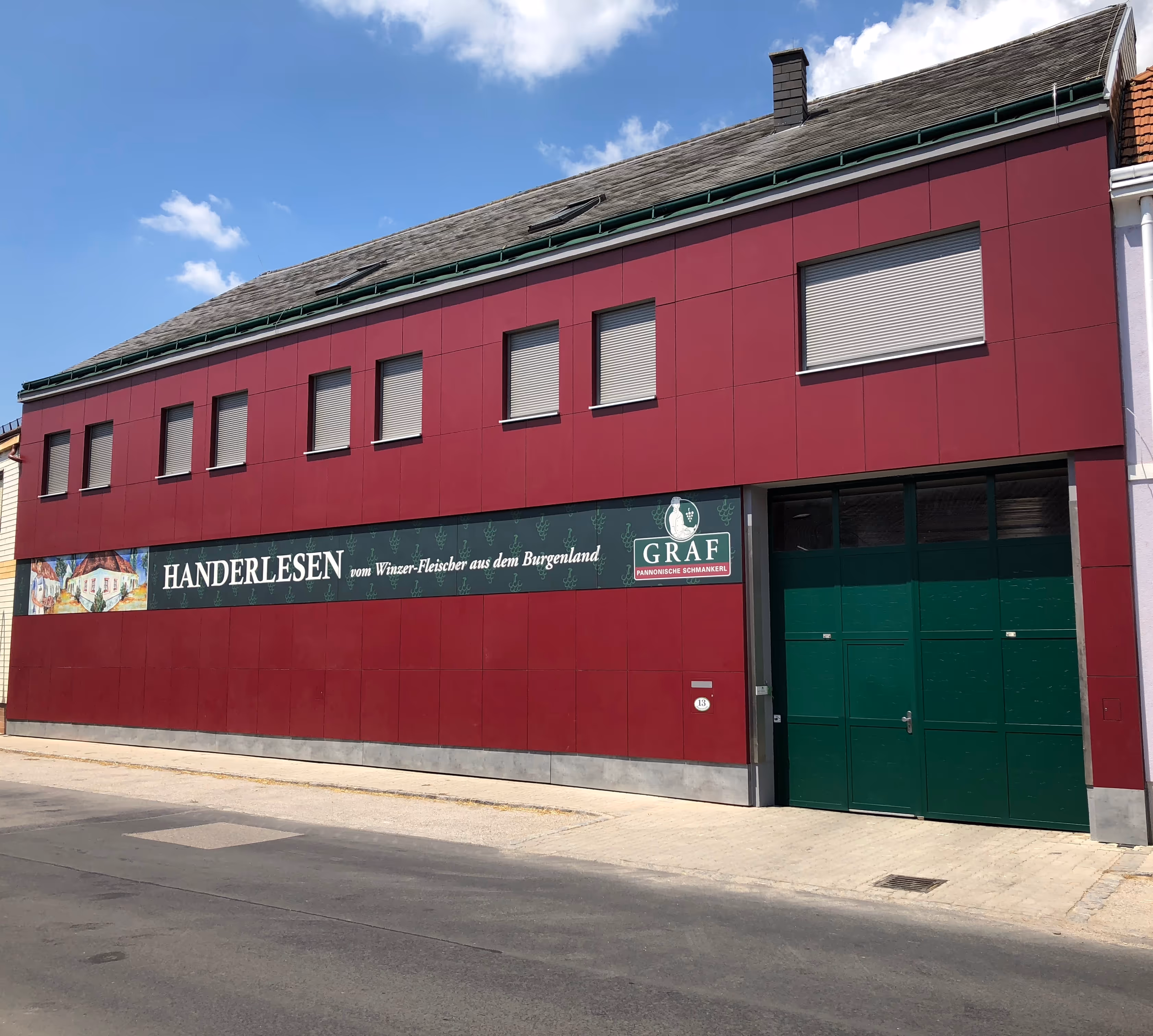 Building with maroon facade, several small closed windows, a large green garage door, and a sign reading 'HANDERLESEN vom Winzer-Fleischer aus dem Burgenland GRAF Pannonische Schmankerl'.