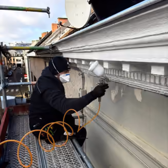Person wearing a mask and black clothes spray painting white trim on a building facade from scaffolding.