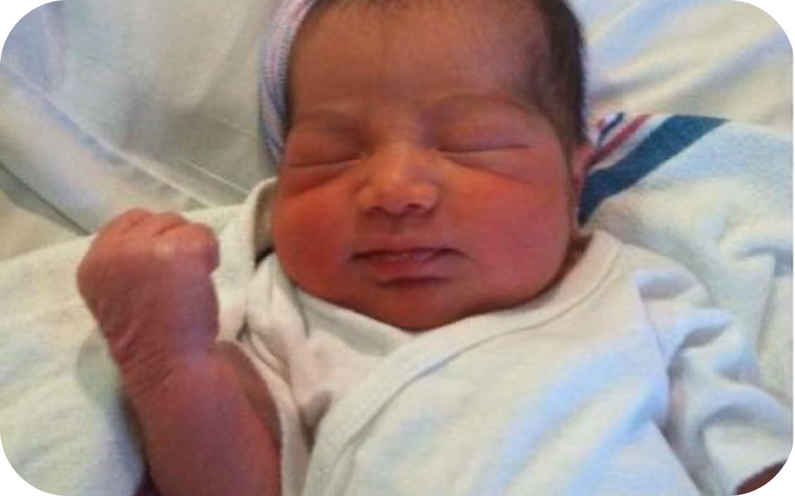 Newborn baby sleeping with eyes closed and one fist raised, wrapped in a white blanket.
