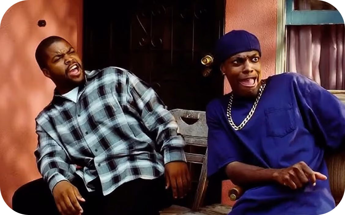 Two men sitting outside on chairs, one in a black and white plaid shirt leaning back with a surprised expression, the other in a blue shirt and cap pointing with a shocked face.