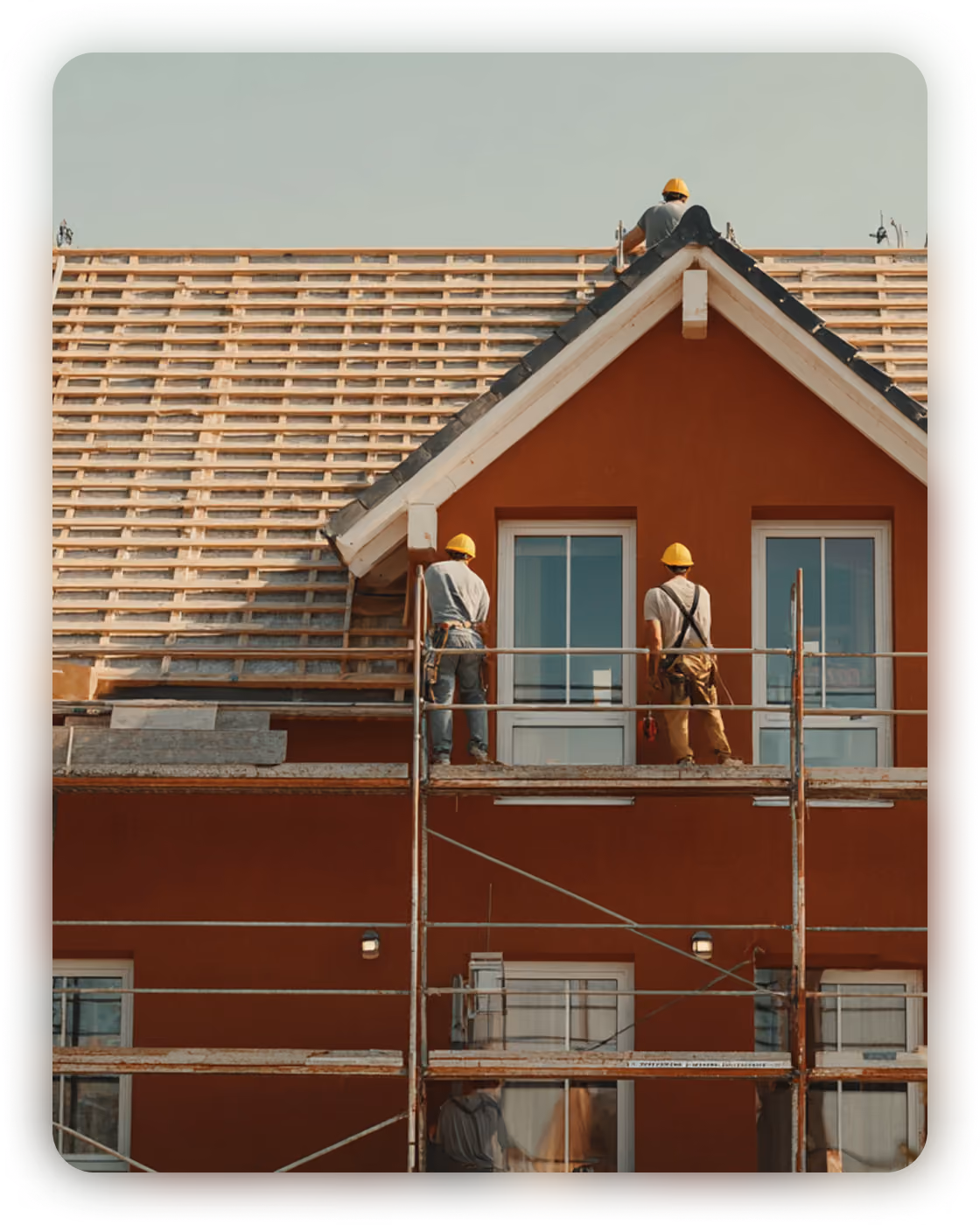 Three construction workers wearing yellow helmets working on scaffolding to install roofing on a red house with large windows.