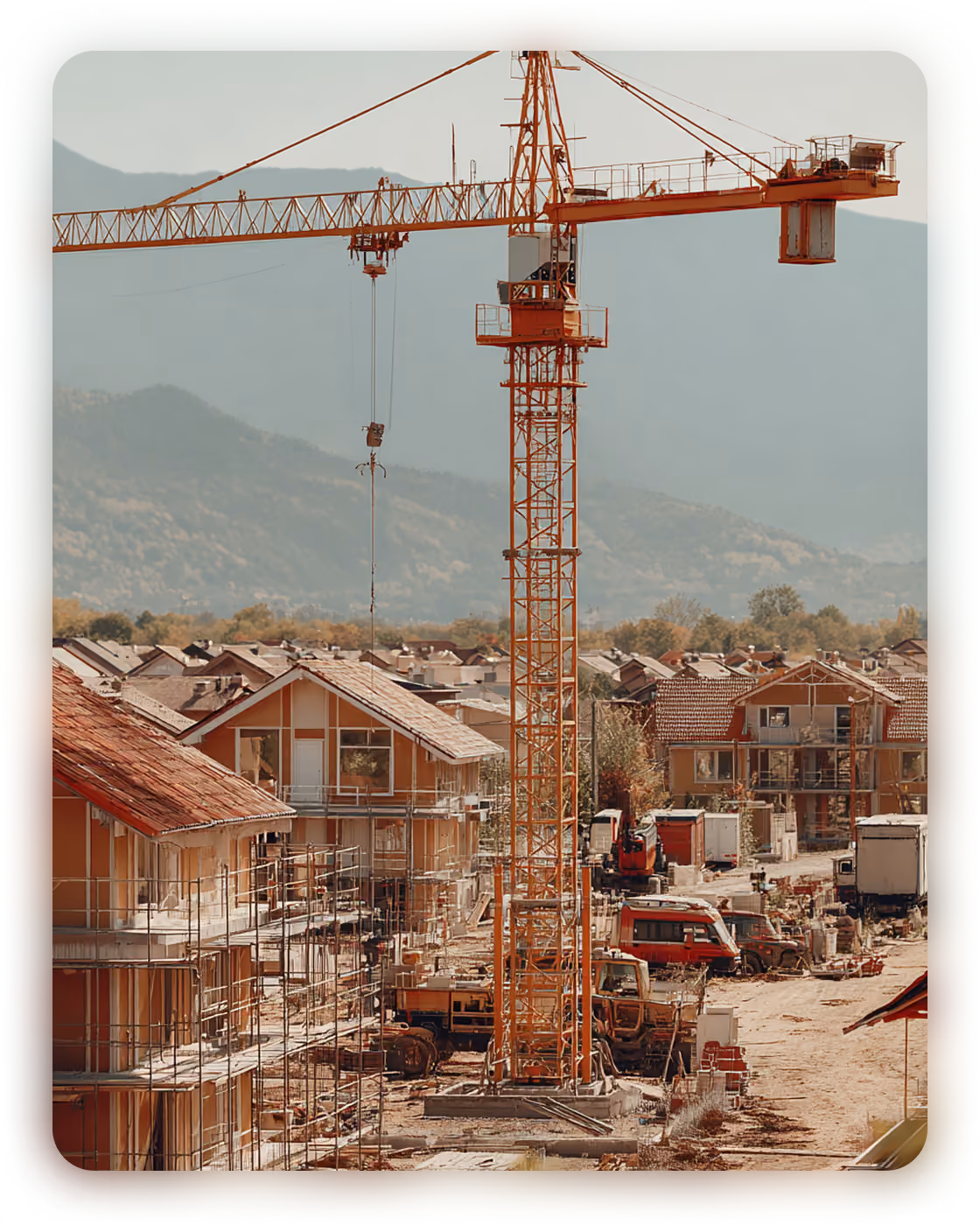 Construction site with a tall yellow crane, scaffolding on partially built houses, and construction vehicles, set against a mountainous background.