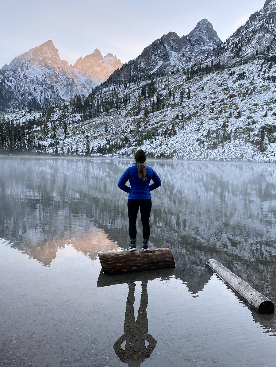 Girl standing on a log on a mirrored lake looking at the snowy mountains