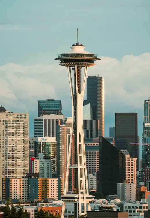 The Space Needle, a tall observation tower in Seattle, stands prominently against a backdrop of various modern skyscrapers and buildings under a cloudy sky.