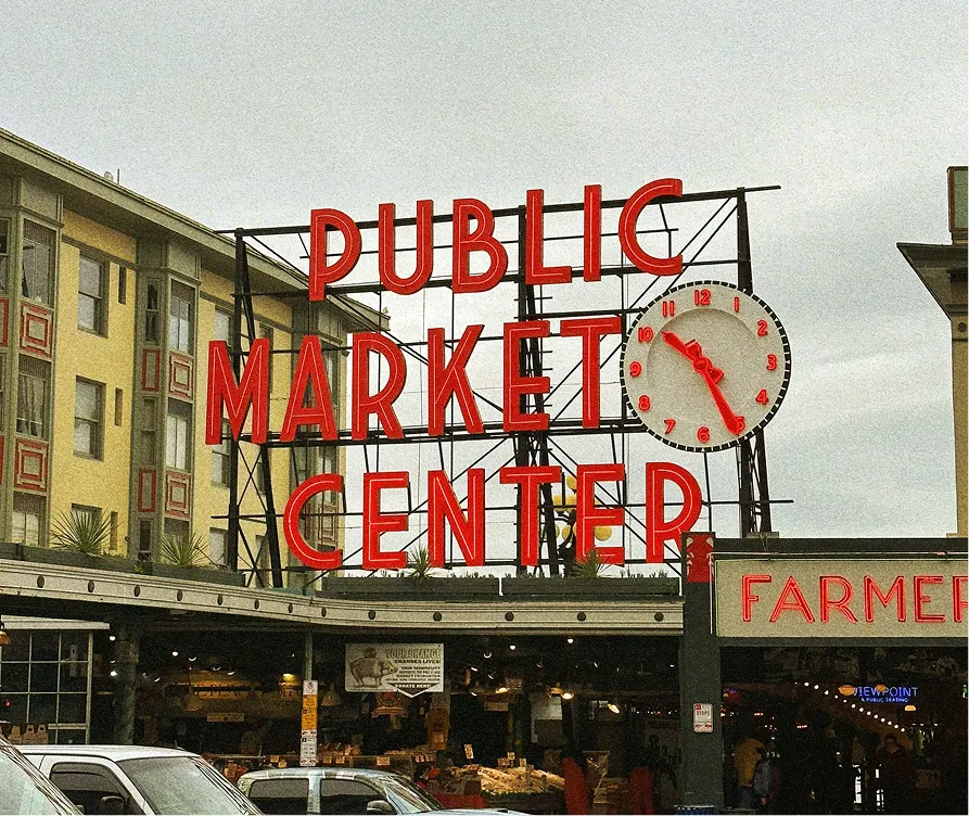 Large red neon sign reading 'PUBLIC MARKET CENTER' with a clock showing 12:26, mounted above a busy market entrance and cars parked below.