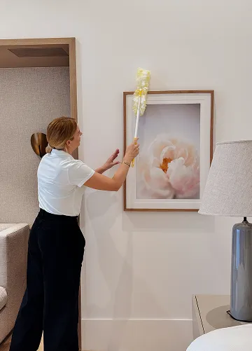 A woman dusting a framed flower photograph on a beige wall using a yellow duster in a modern room with a lamp and sofa.