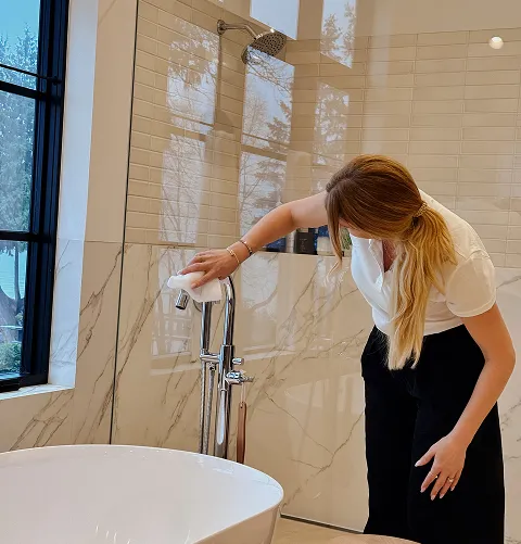 A woman with long blonde hair cleaning a chrome bathtub faucet in a modern bathroom with marble walls and a large window.