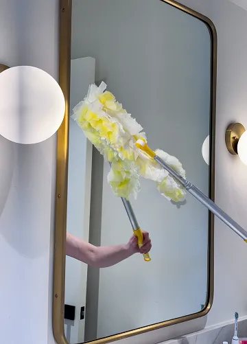 Hand holding two yellow and white dusters in front of a rectangular mirror with a gold frame, with white spherical wall lights reflected on both sides of the mirror.