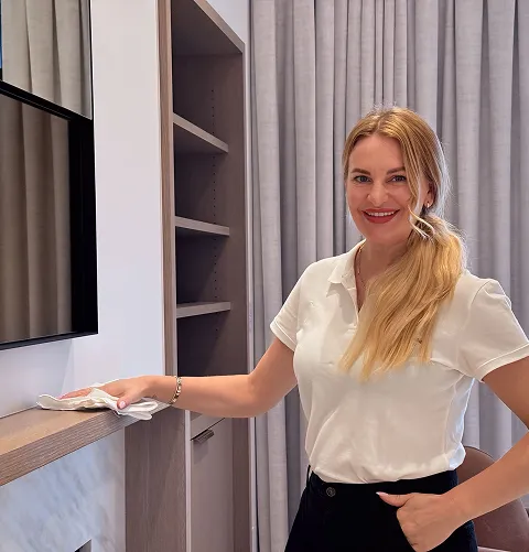 Smiling woman with long blonde hair in a white polo shirt cleaning a wooden shelf near a television in a modern interior space.