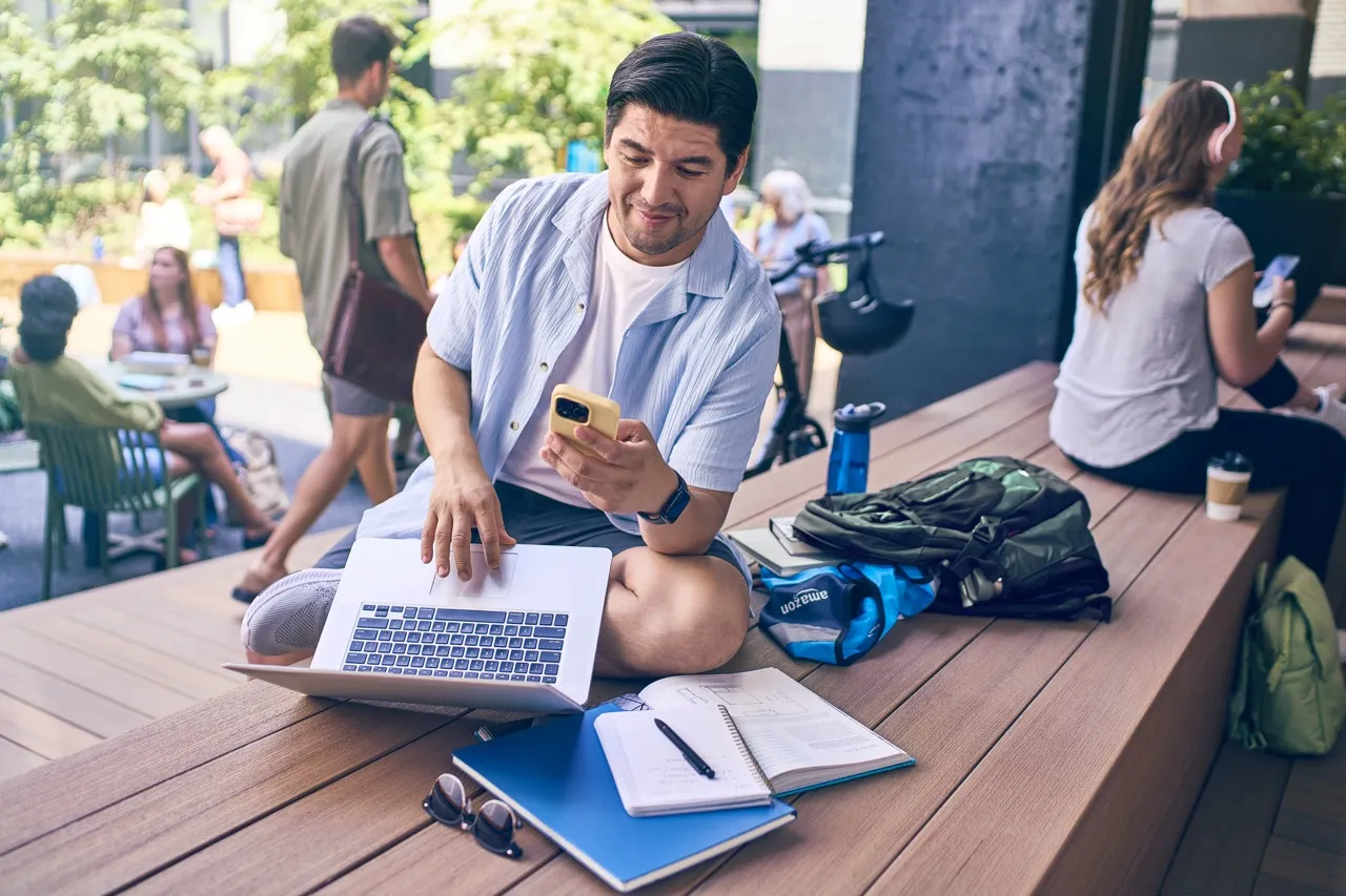 A man sits on a large community bench and looks down at a phone he holds in one hand. The other hand types on a laptop that sits on his lap.
