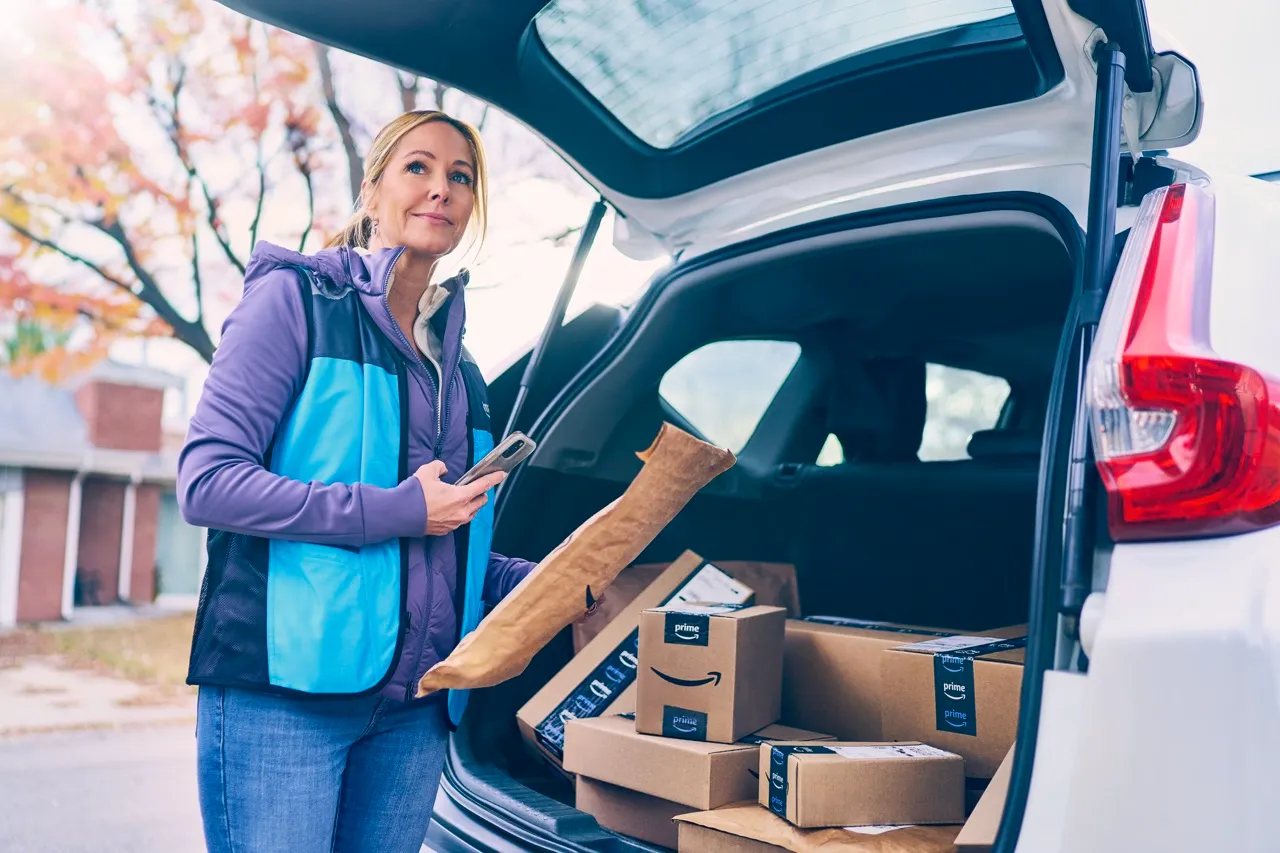 A woman stands behind the open trunk of her SUV and scans a package with her phone. She wears a blue Amazon vest, and in the back of the trunk are half a dozen more packages.